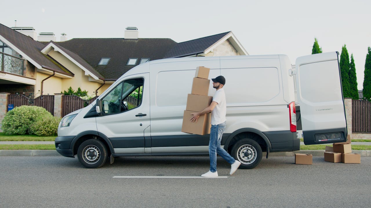 Man unloading cardboard boxes from a delivery van in a suburban neighborhood street.